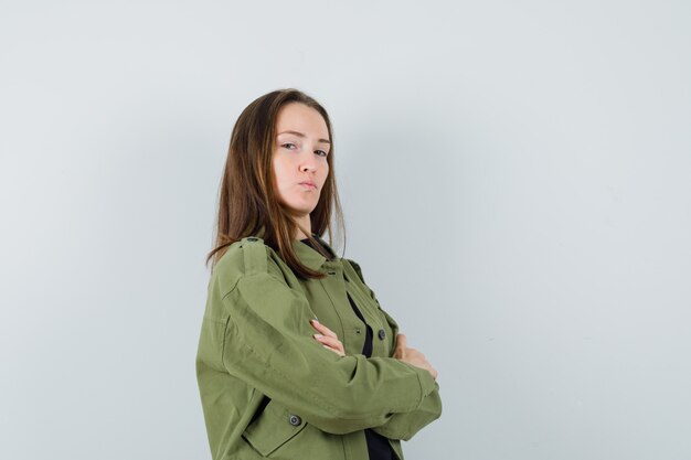 Young woman standing with crossed arms while looking aside in green jacket,black shirt and looking boastful , front view.