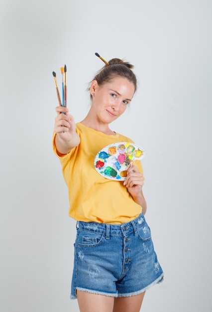 Молодая женщина стоит с инструментами для рисования в желтой футболке, джинсовых шортах и выглядит веселой