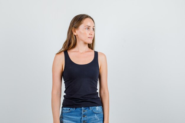 Young woman staring at side in singlet, shorts and looking pensive. front view.