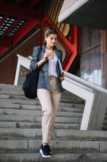 Young woman stepping up-down stair holding disposable coffee cup and digital tablet