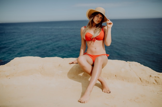 Young woman in straw hat and bikini sitting on the end of rock