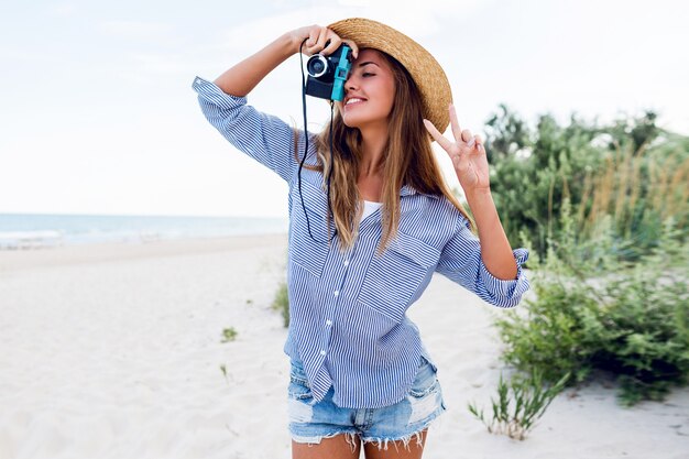 Young woman in straw hat making picture with retro camera on the beach