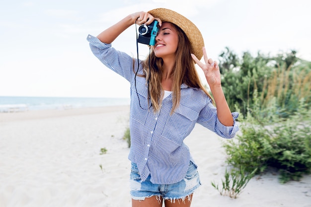 Молодая женщина в соломенной шляпе делает снимок с ретро камерой на пляже