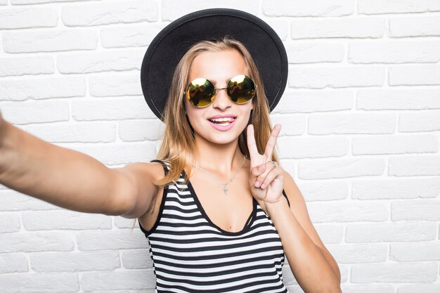 Молодая женщина в соломенной шляпе и солнцезащитных очках делает селфи смартфон с жестом мира над белой кирпичной стеной офиса