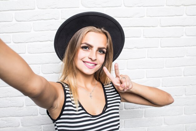 Young woman in straw hat and sunglasses taking selfie smart phone with peace gesture over white brick office wall