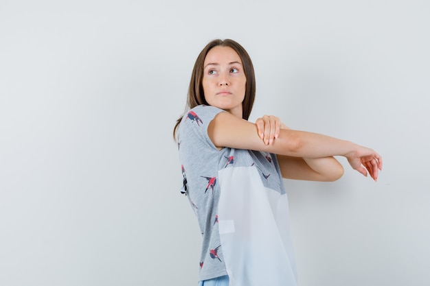 Free photo young woman stretching arm while looking back in t-shirt, skirt and looking curious , front view.