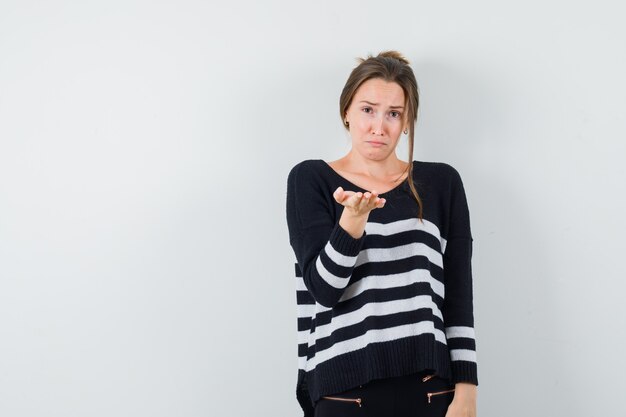 Young woman stretching hand as receiving something in striped knitwear and black pants and looking sad