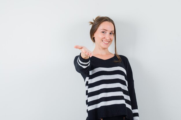 Young woman stretching hand toward front in striped knitwear and black pants and looking happy