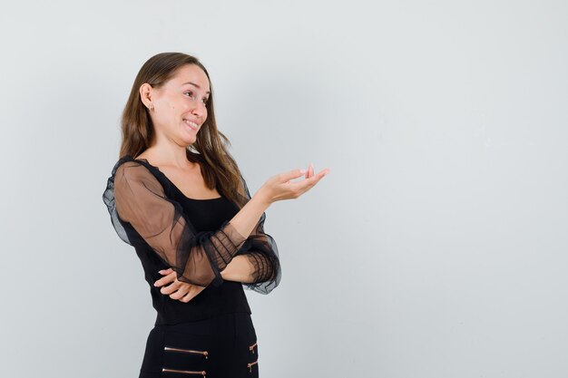 Young woman stretching hand toward right and smiling in black blouse and black pants and looking happy 