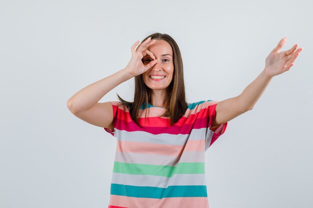 若い女性は、Tシャツを着て、目がOKのサインで手を伸ばし、陽気に見えます。正面図。