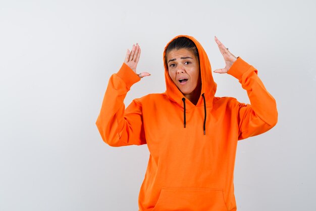 Young woman stretching hands as holding something in orange hoodie and looking surprised