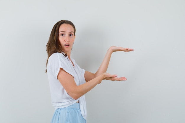 Free photo young woman stretching hands as presenting something in white blouse and light blue skirt and looking surprised