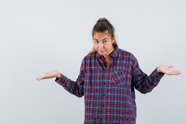 Young woman stretching hands in questioning manner in checked shirt and looking puzzled