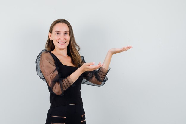 Young woman stretching hands toward right as presenting something and smiling in black blouse and black pants and looking happy. front view.