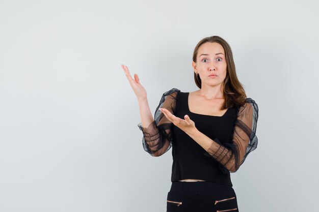 Young woman stretching hands upper left in black blouse and black pants and looking surprised. front view.