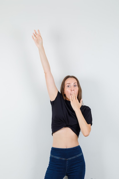 Young woman stretching her arms while covering her mouth in black blouse and looking weird