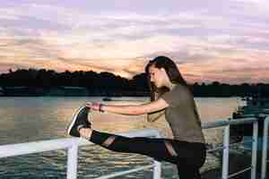 Foto gratuita giovane donna che allunga la gamba vicino al fiume al tramonto