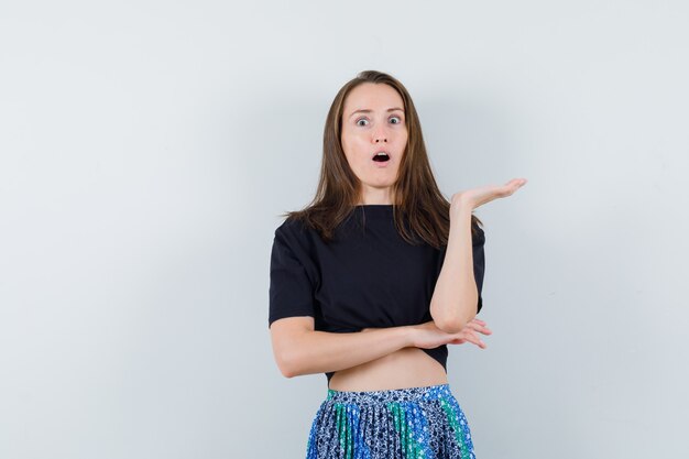 Young woman stretching one hand as holding something in black t-shirt and blue skirt and looking surprised