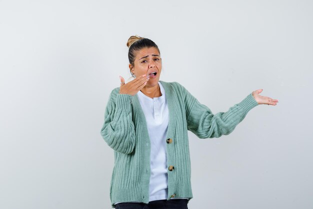 Young woman stretching one hand as holding something and trying to cover mouth with hand in white t-shirt and mint green cardigan and looking surprised