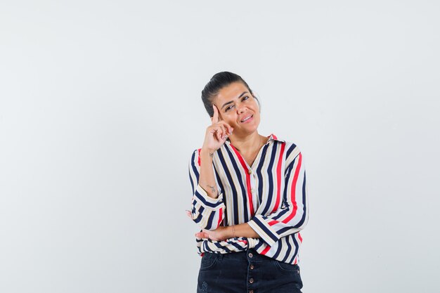 Young woman in striped blouse leaning cheek on hand and standing in listening pose and looking optimistic