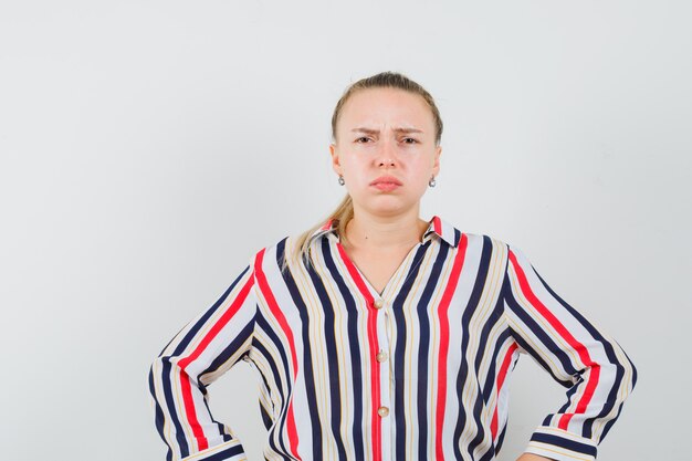 Young woman in striped blouse putting her hands on hips and looking confused