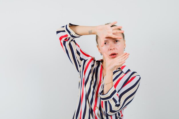 Young woman in striped blouse showing camera hand gesture and looking surprised