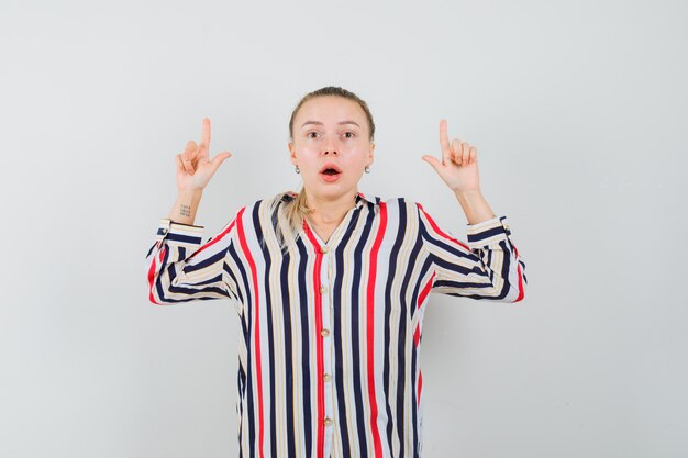 Young woman in striped blouse showing loser gestures with both hands and looking surprised