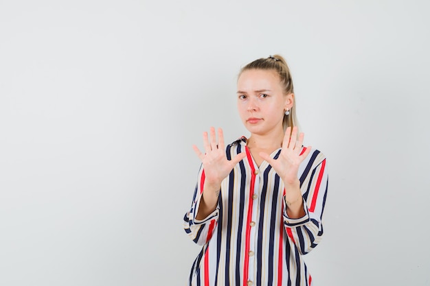 Young woman in striped blouse showing stop gestures with both hands and looking frightened