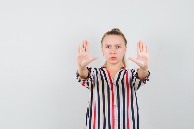 Young woman in striped blouse showing stop gestures with both hands and looking serious