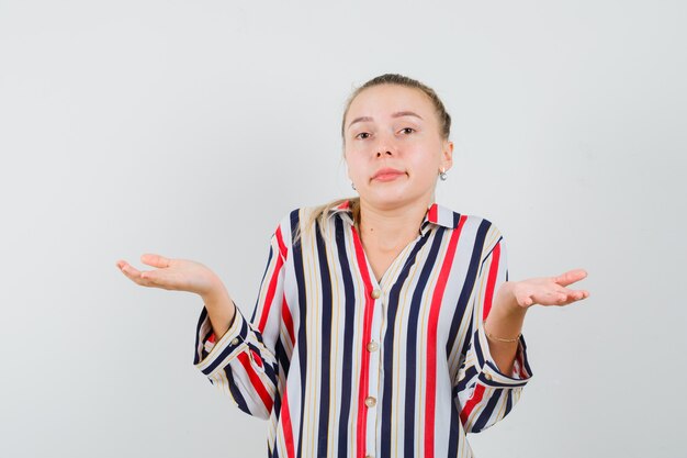 Young woman in striped blouse shrugging her shoulders and raising her hands and looking doubtful