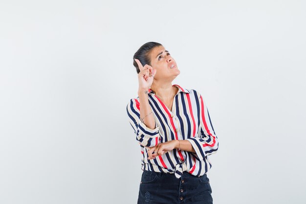 Young woman in striped blouse standing in thinking pose and raising finger in eureka gesture and looking pensive