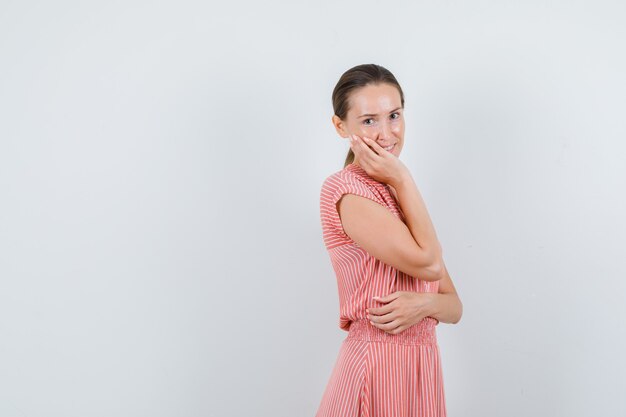 Young woman in striped dress holding fingers on cheek and looking cheerful , front view.