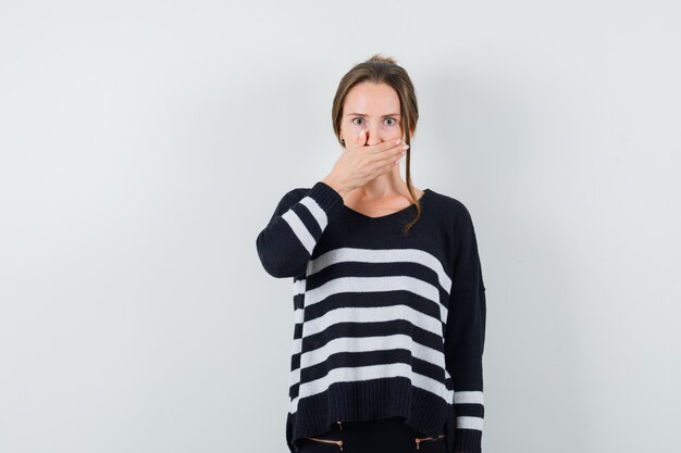 Young woman in striped knitwear and black pants covering mouth with hand and looking shocked, front view.