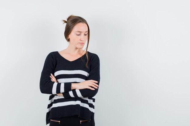 Young woman in striped knitwear and black pants holding arms crossed, looking down and looking pensive