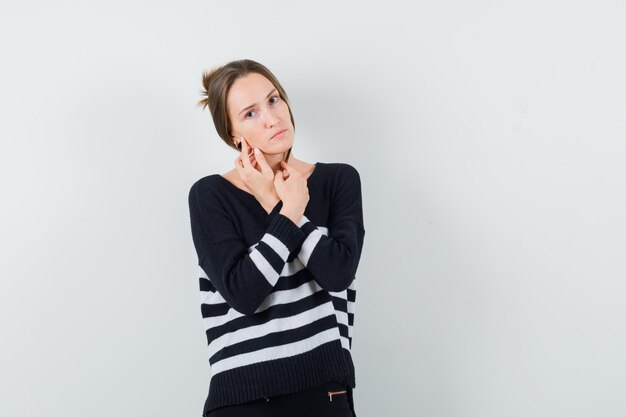 Young woman in striped knitwear and black pants keeping arms crossed and looking happy