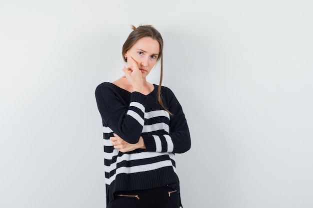 Young woman in striped knitwear and black pants leaning cheek on palm and standing in thinking pose and looking serious