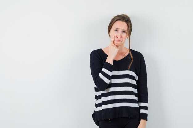 Young woman in striped knitwear and black pants pointing cheek with index finger and looking dismal