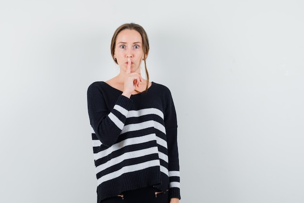 Foto gratuita giovane donna in maglieria a righe e pantaloni neri che mette il dito indice sulla bocca e mostra il gesto di silenzio e sembra seria