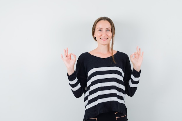 Young woman in striped knitwear and black pants showing ok sign with both hands and looking happy