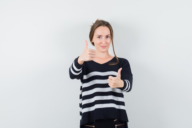 Young woman in striped knitwear and black pants showing thumbs up and looking confident