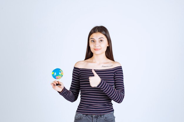 Молодая женщина в полосатой рубашке держит мини-глобус и выглядит взволнованной