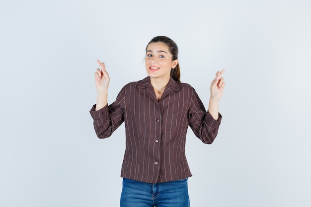 Young woman in striped shirt, jeans keeping fingers crossed and looking happy , front view.