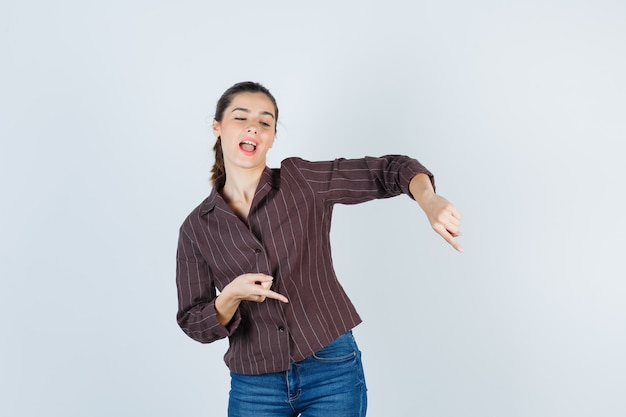 Giovane donna in camicia a righe, jeans rivolti verso il basso con l'indice, tenendo la bocca aperta e guardando felice, vista frontale.