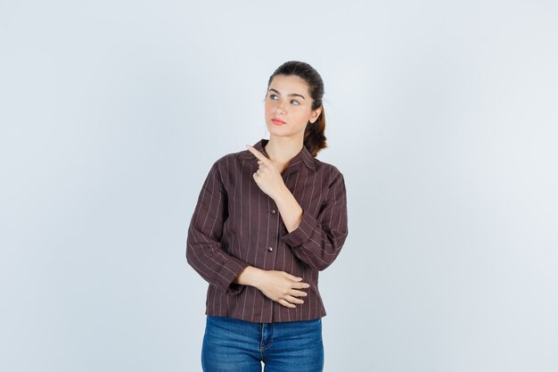 Young woman in striped shirt, jeans pointing left with index finger and looking focused , front view.