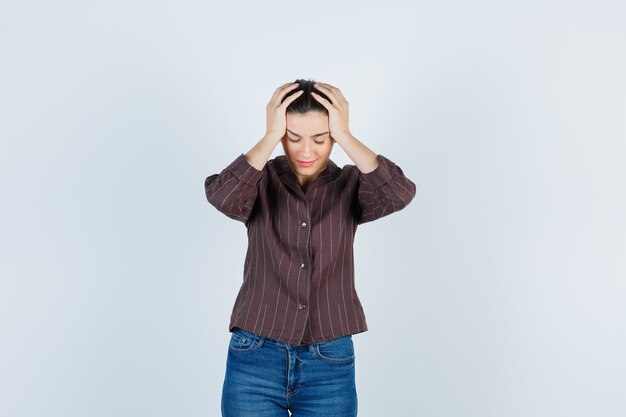 Young woman in striped shirt, jeans with hands on head and looking exhausted , front view.