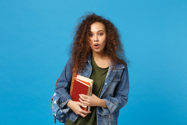 Молодая женщина-студент в джинсовой одежде и рюкзаке держит книги, изолированные на синей стене