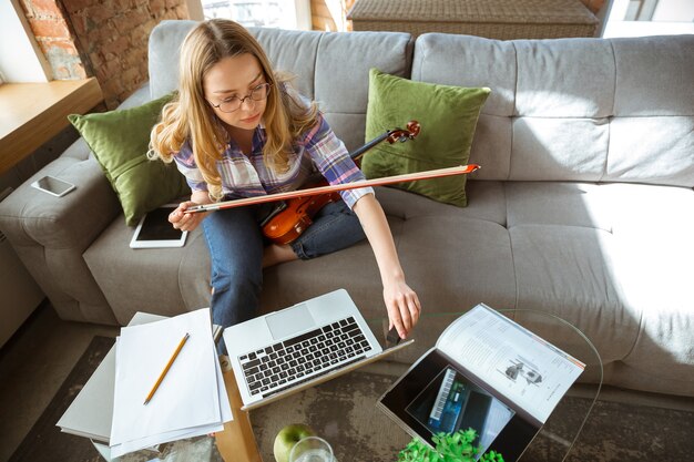 Young woman studying at home during online courses or free information by herself. Becomes musician, violinist while isolated, quarantine against coronavirus spreading. Using laptop, smartphone.