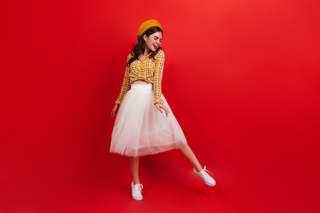 Young woman in stylish orange hat and bright blouse is dancing on red wall. Girl in white skirt and sneakers smiles.