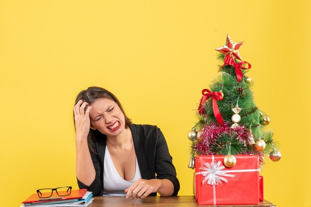 Young woman suffering from headache sitting at a table in suit near decorated Christmas tree at office on yellow 