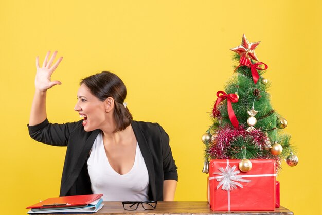 Young woman in suit calling someone near decorated Christmas tree at office on the right side on yellow 